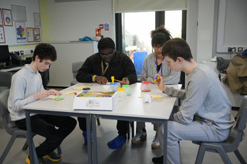 Teacher guiding three students painting dinosaur crafts using adaptive tools with non-spill paint pots and easy-grip brushes at a classroom table.