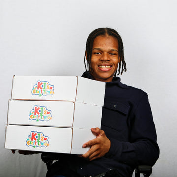 Khamani Edwards, founder of KJ's Craft Time, holding stacked boxes with 'KJ's Craft Time' logo branding against a plain background