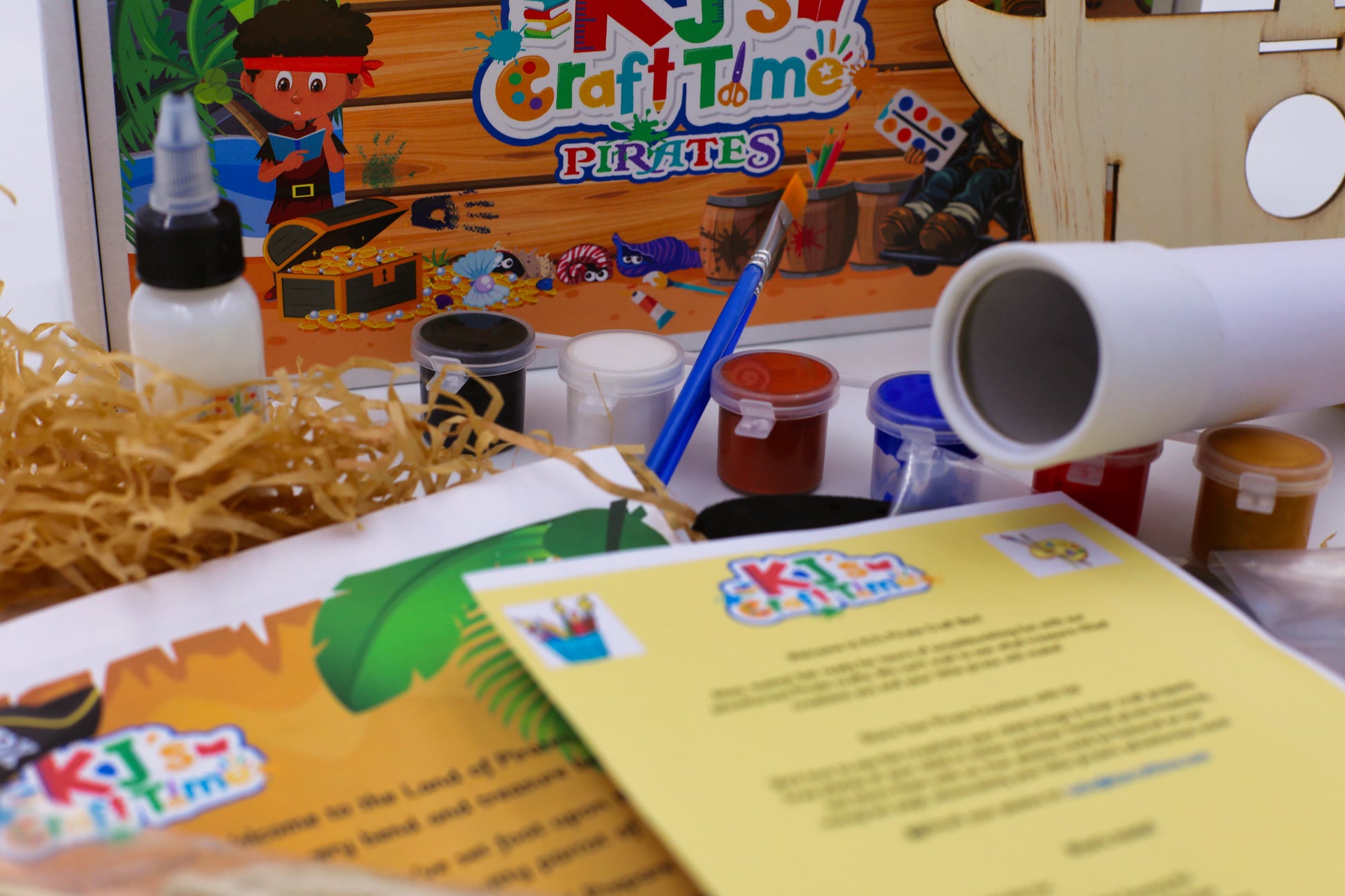 Close-up of pirate craft kit materials including paints, paintbrush, PVA glue, telescope craft and pirate-themed activity sheets.